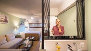A hotel room with a bed, a sofa, a coffee table, a mirror, and a bathroom with a sink and mirror. - Pols Studio - Project Kalamata Mediterranean Suites (Kalamata)