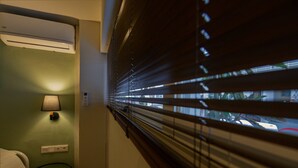 A hotel room with a bed, a nightstand with a lamp, a small table with a card, and a window with blinds. - Pols Studio - Project Kalamata Mediterranean Suites (Kalamata)