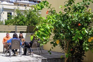 Un groupe de personnes est assis à une table à l’extérieur, entouré de chaises. On aperçoit une plante en pot avec des feuilles vertes et quelques fleurs rouges, ainsi qu’un bâtiment avec des balcons en arrière-plan.