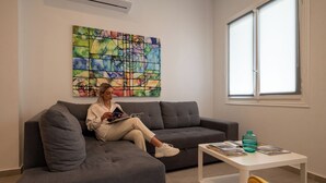 Una persona sentada en un sofá gris en una habitación con un cuadro abstracto de colores en la pared, una ventana y una pequeña mesa blanca con revistas y una botella de agua.