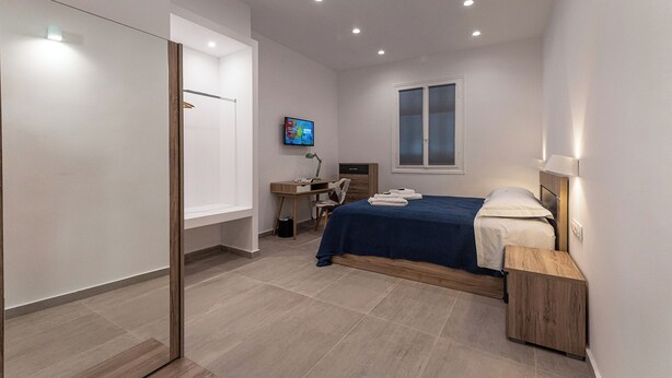 Un dormitorio moderno con cama, mesita de noche, escritorio, silla y televisión en la pared.