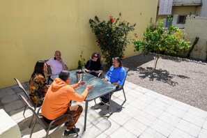 Un groupe de personnes est assis autour d’une table à l’extérieur, avec un mur jaune et des plantes en arrière-plan.