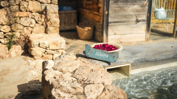 A stone wall, a wooden door, and a small wooden box labeled "Sauna" with pink flowers. - Es Cucons Rural Hotel (Sant Antoni de Portmany)