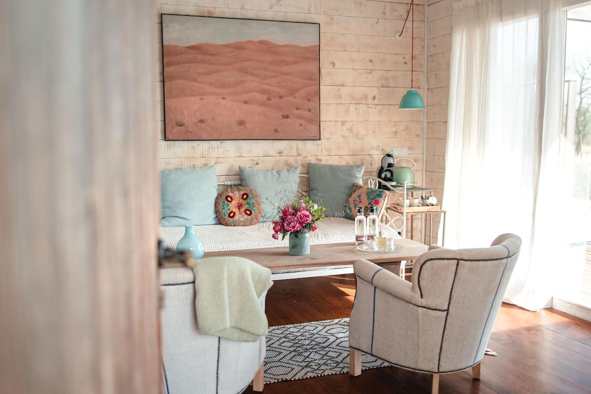 A cozy living room with a wooden wall, a large painting, a sofa with patterned cushions, a small table with decorative items, and a chair.