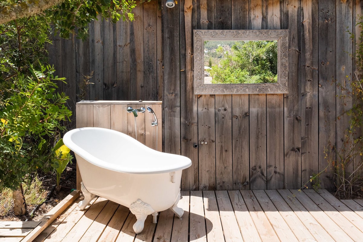 A wooden deck with a white freestanding bathtub and a window with a view of greenery.