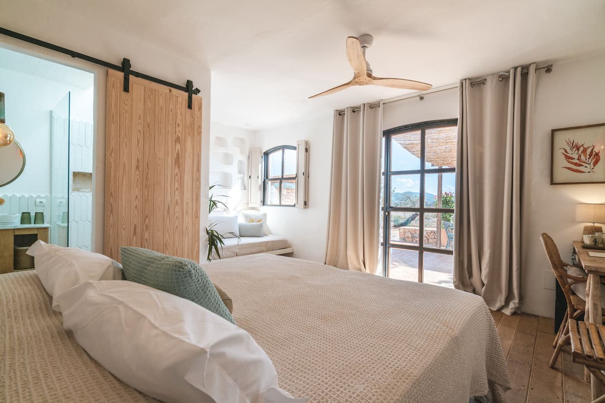 A bedroom with two beds, a wooden headboard, a ceiling fan, a desk with a chair, and a view of the outdoors.