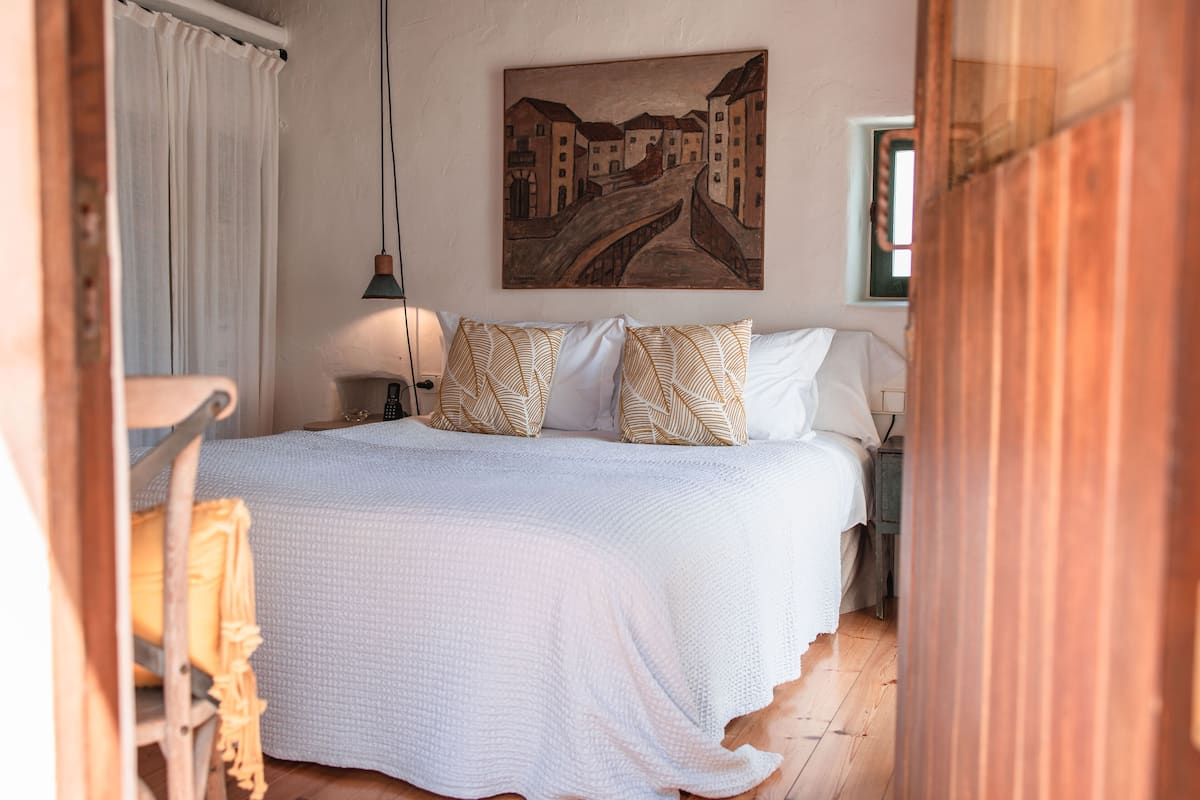 A bedroom with a large bed, a painting on the wall, and a window.