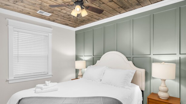 A bedroom with a wooden ceiling, a bed with white bedding, two bedside lamps, and a window with blinds. - Downtown Access | Spacious | Family Space (Starkville)