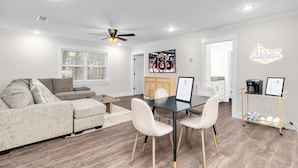 A modern living room with a sofa, a dining table with chairs, a ceiling fan, and a kitchen area with a coffee machine and framed certificates. - Downtown Access | Spacious | Family Space (Starkville)