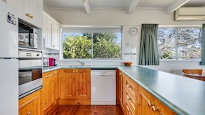 A modern kitchen with wooden cabinets, a dishwasher, and a large window with a view of trees. - Beach House On Booth ~ The Quintessential Beach House for the Family Get-away (Narrawallee)