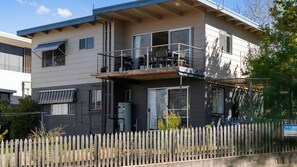 A two-story building with a balcony, a fenced yard, and a sign indicating holiday accommodation. - Beach House On Booth ~ The Quintessential Beach House for the Family Get-away (Narrawallee)