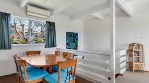 A dining area with a wooden table and chairs, a window with curtains, a wall-mounted air conditioner, and a white railing. - Beach House On Booth ~ The Quintessential Beach House for the Family Get-away (Narrawallee)