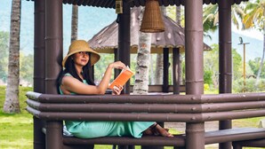 A person sitting on a bench under a pavilion with a thatched roof, reading a book. - Avana Resort (Mettupalayam)