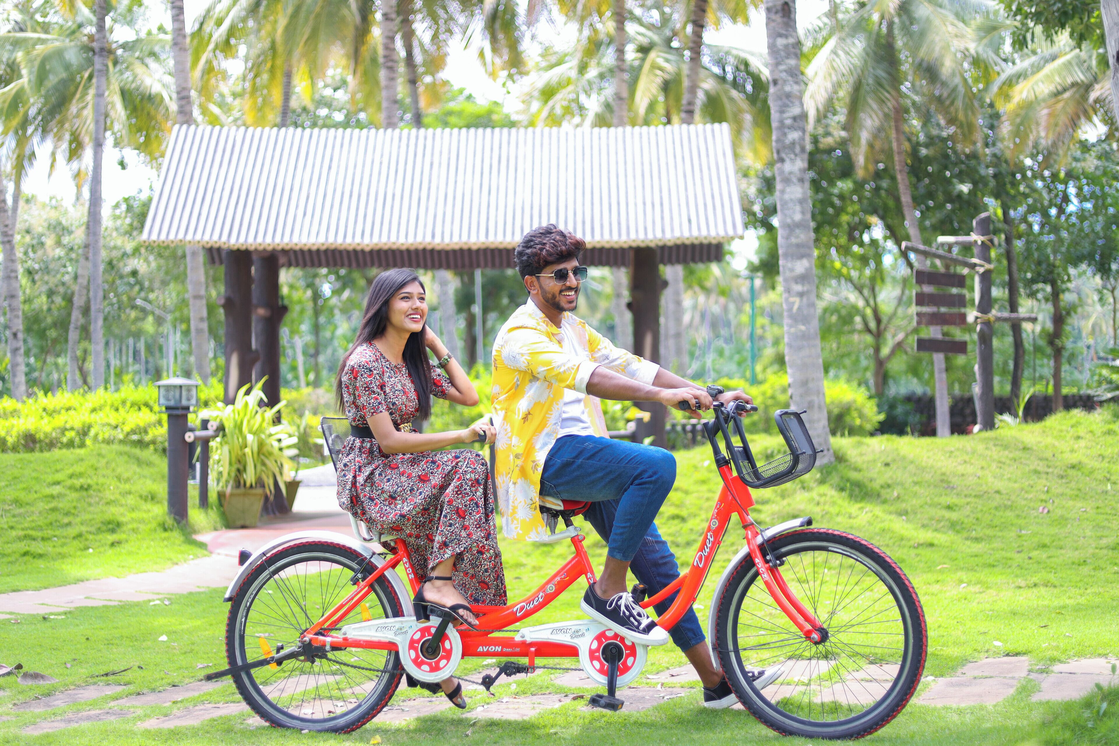 Una pareja en una bicicleta roja en un jardín exuberante con un pabellón al fondo.