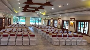 A large hall with rows of chairs, each with a white cover and red sashes, and large windows along the walls. - Avana Resort (Mettupalayam)