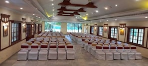 Un salón amplio con filas de sillas, cada una cubierta con una funda blanca y listones rojos, y grandes ventanales a lo largo de las paredes.