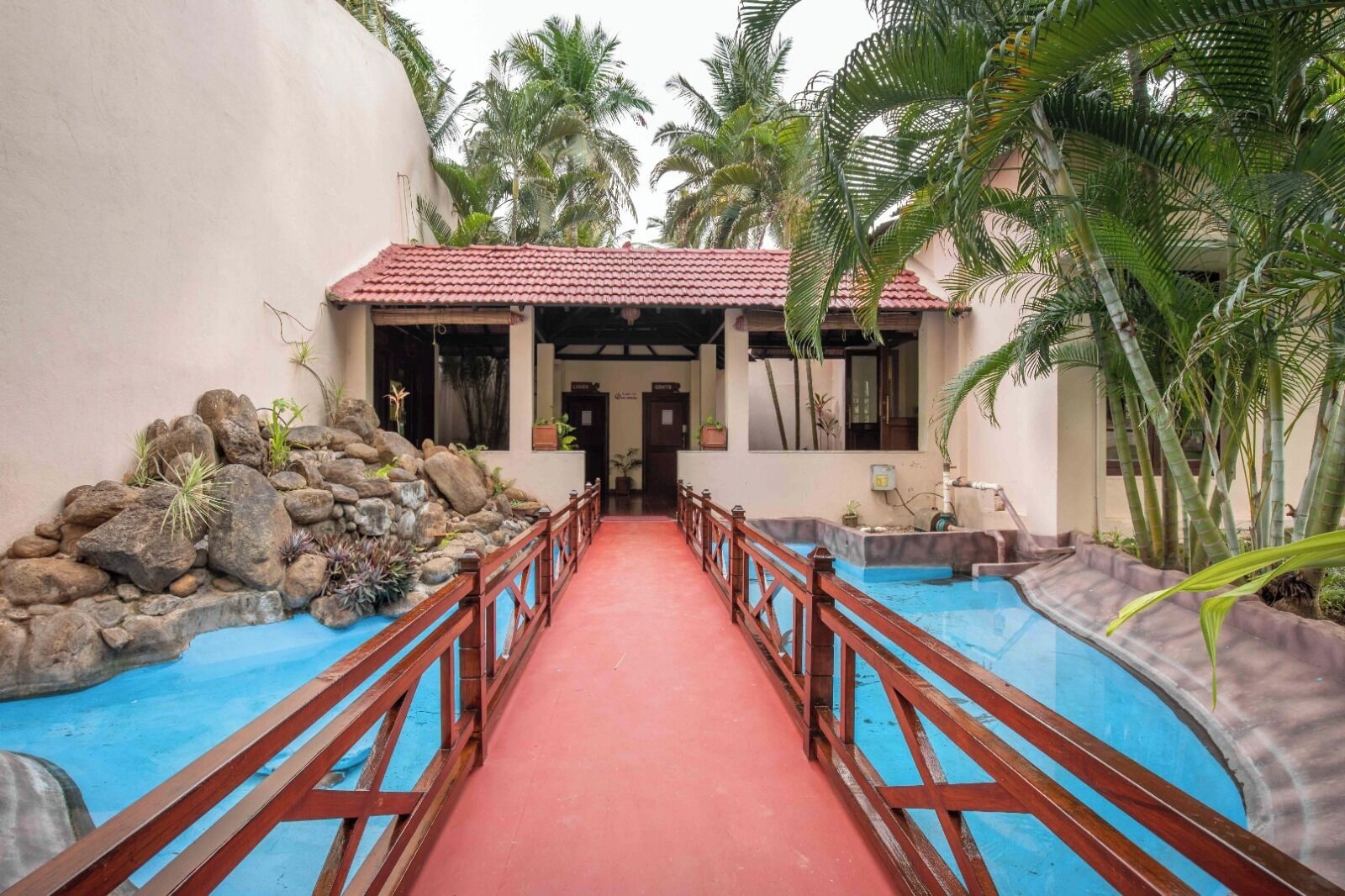 Uma passagem leva a um edifício com telhado vermelho, cercado por uma piscina e palmeiras.