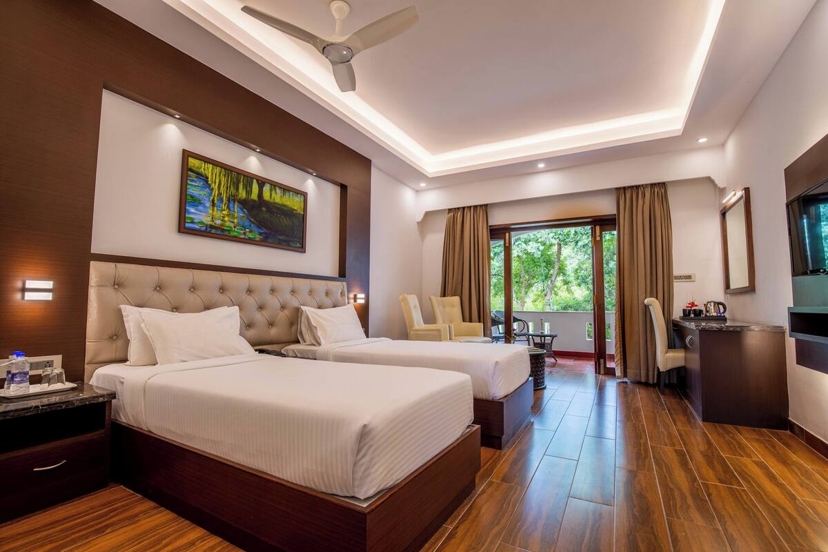 A hotel room with two beds, a desk, a chair, and a view of greenery outside.