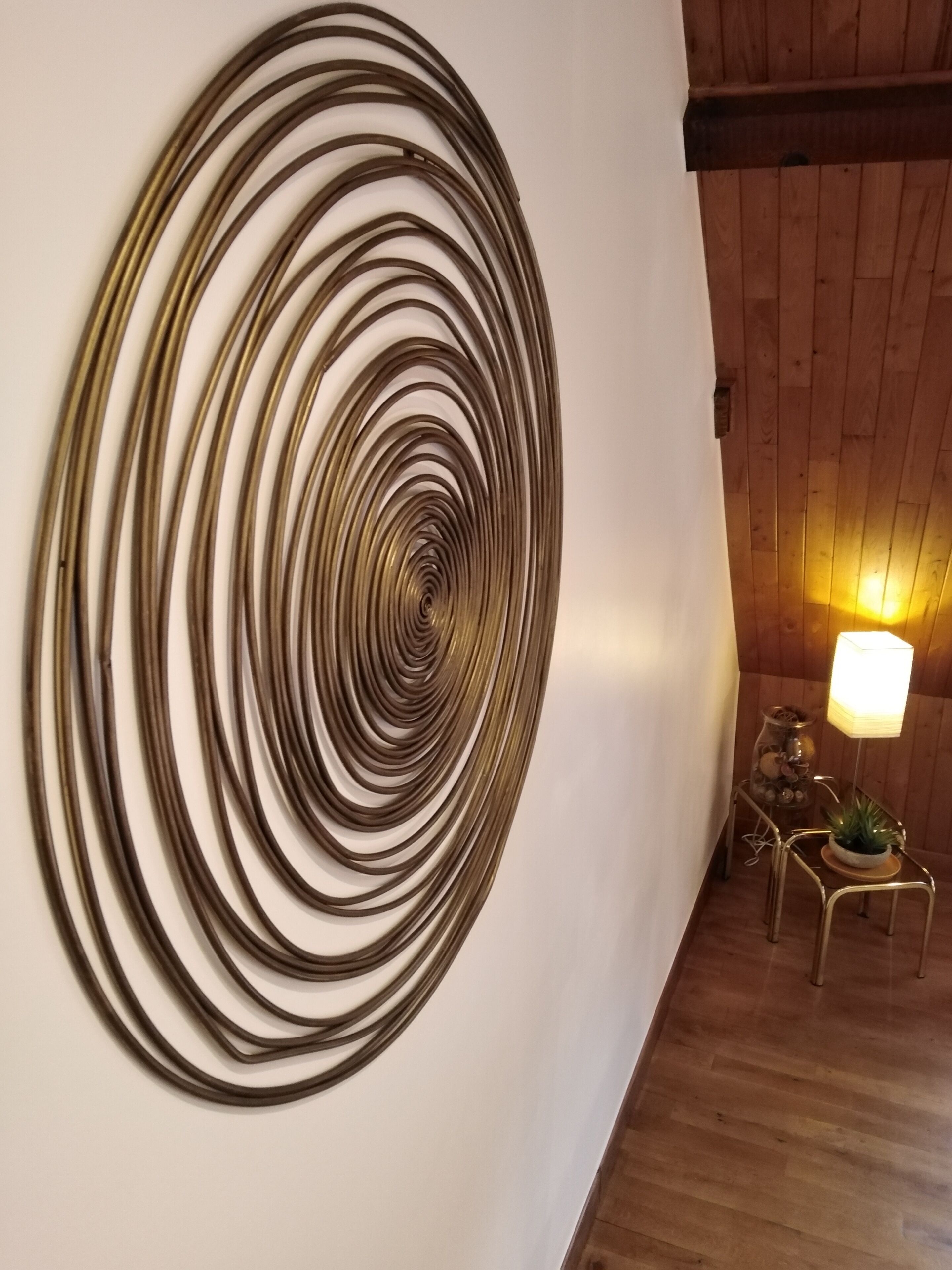 Un'installazione artistica a spirale su una parete in una stanza con pavimento in legno e una lampada.