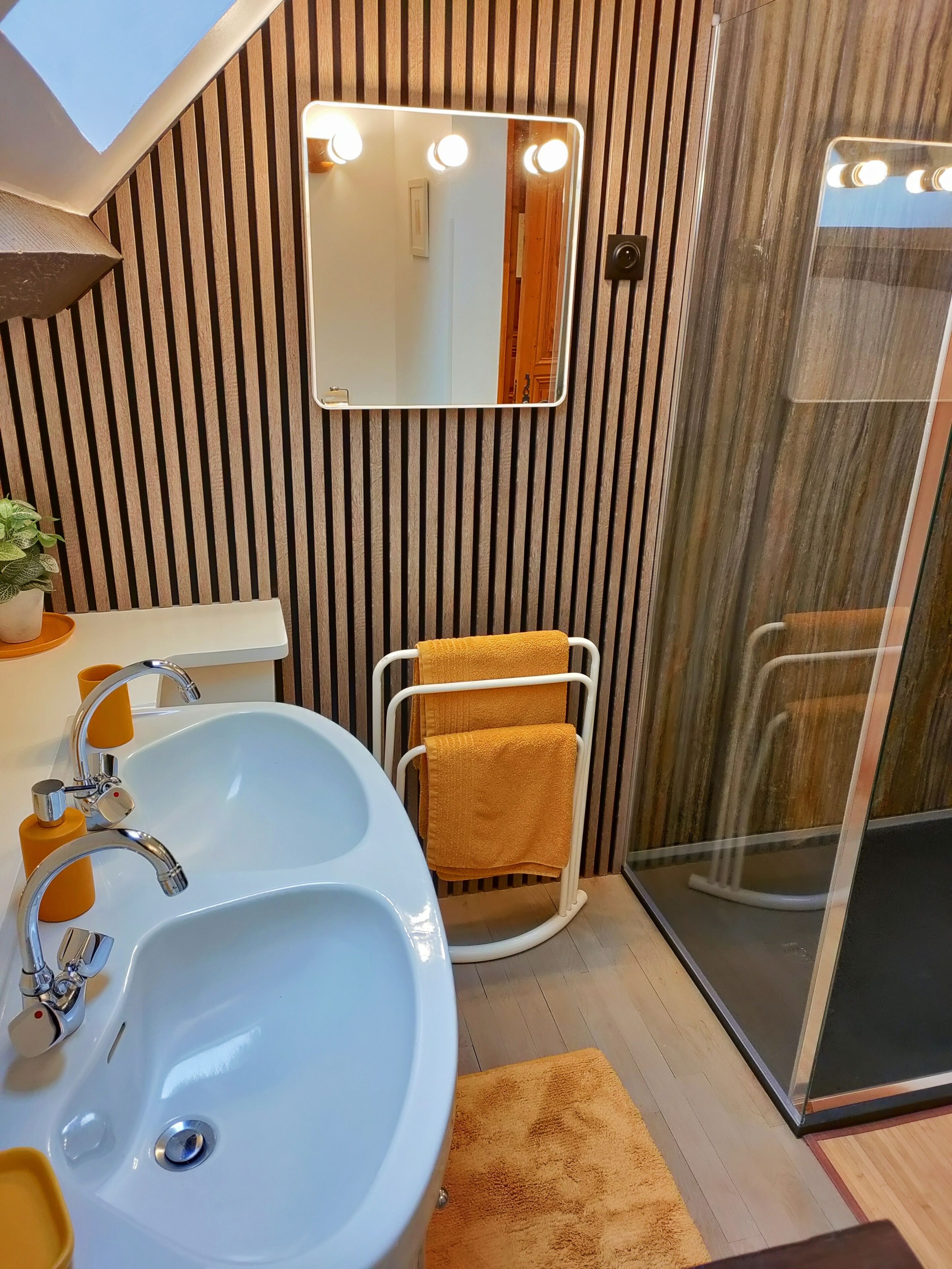 Un baño moderno con un lavamanos blanco, grifos cromados, un perchero con una toalla amarilla, un espejo con luces y una cabina de ducha de vidrio.