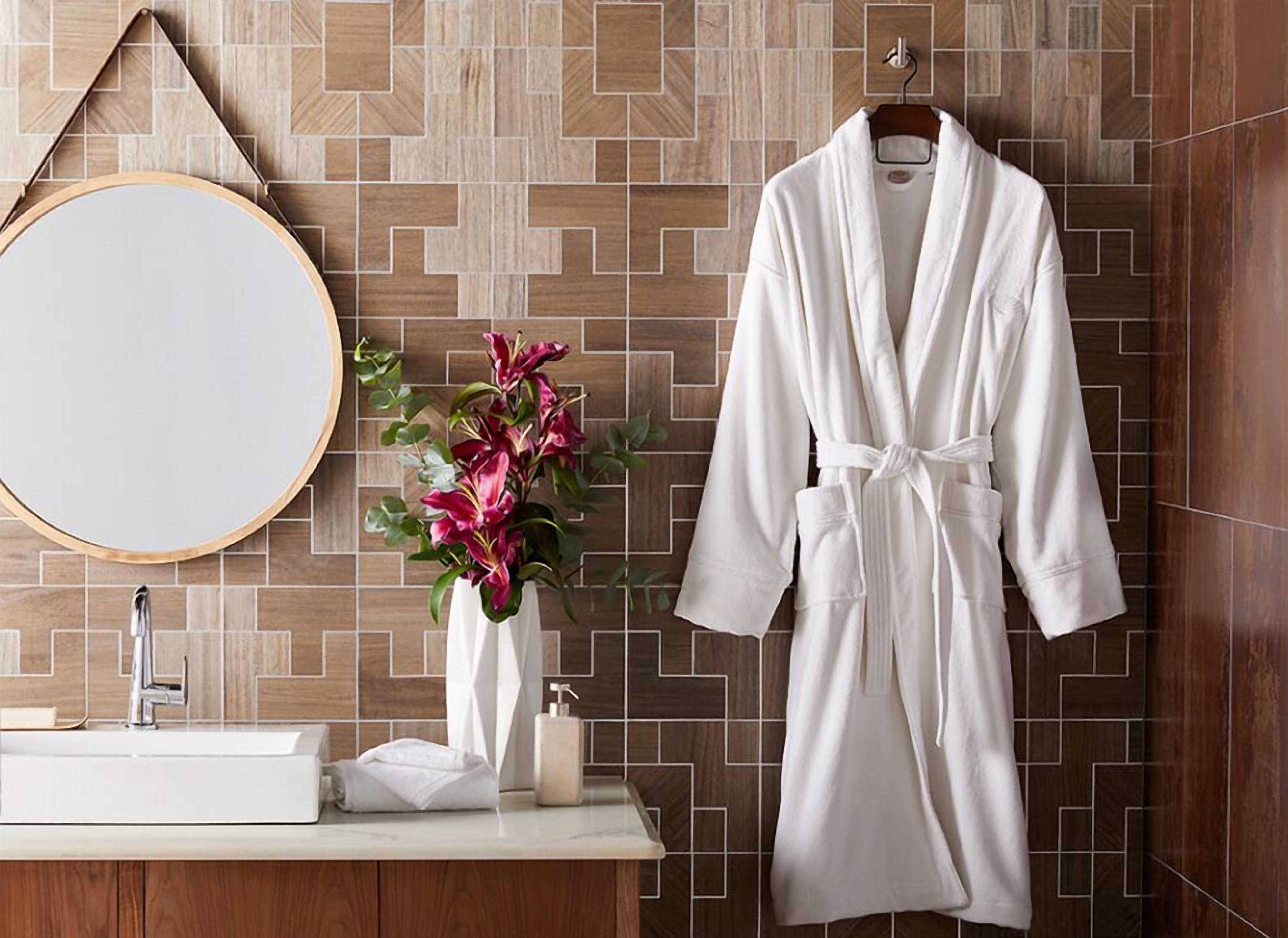 Une salle de bain avec un plancher en bois, un miroir rond, un lavabo blanc, un peignoir blanc accroché à un porte-serviettes et un vase rempli de fleurs roses.