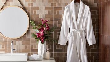 Une salle de bain avec un plancher en bois, un miroir rond, un lavabo blanc, un peignoir blanc accroché à un porte-serviettes et un vase rempli de fleurs roses.