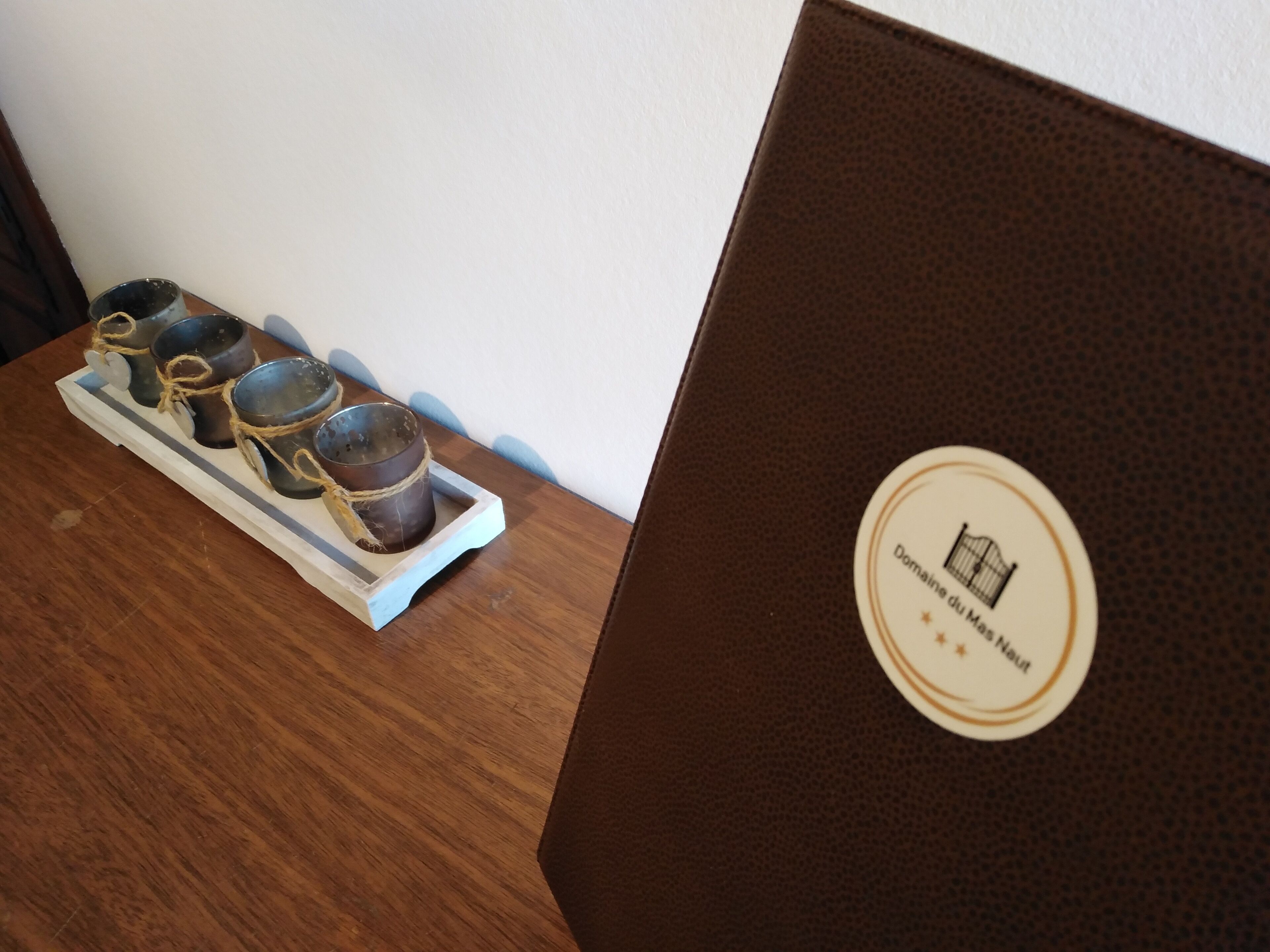 A wooden table with a tray holding four candles tied with twine. A brown leather-bound book or folder with a logo is on the right.