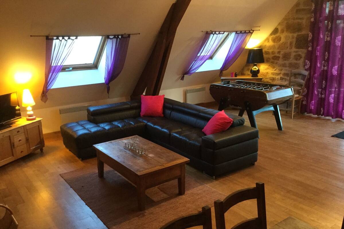 Un salon chaleureux avec un canapé d’angle, une table basse en bois, une table de baby-foot et un téléviseur posé sur un meuble.