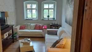Ett mysigt vardagsrum med en soffa, ett litet bord och en tv. Det finns två fönster med utsikt över grönområden utanför.