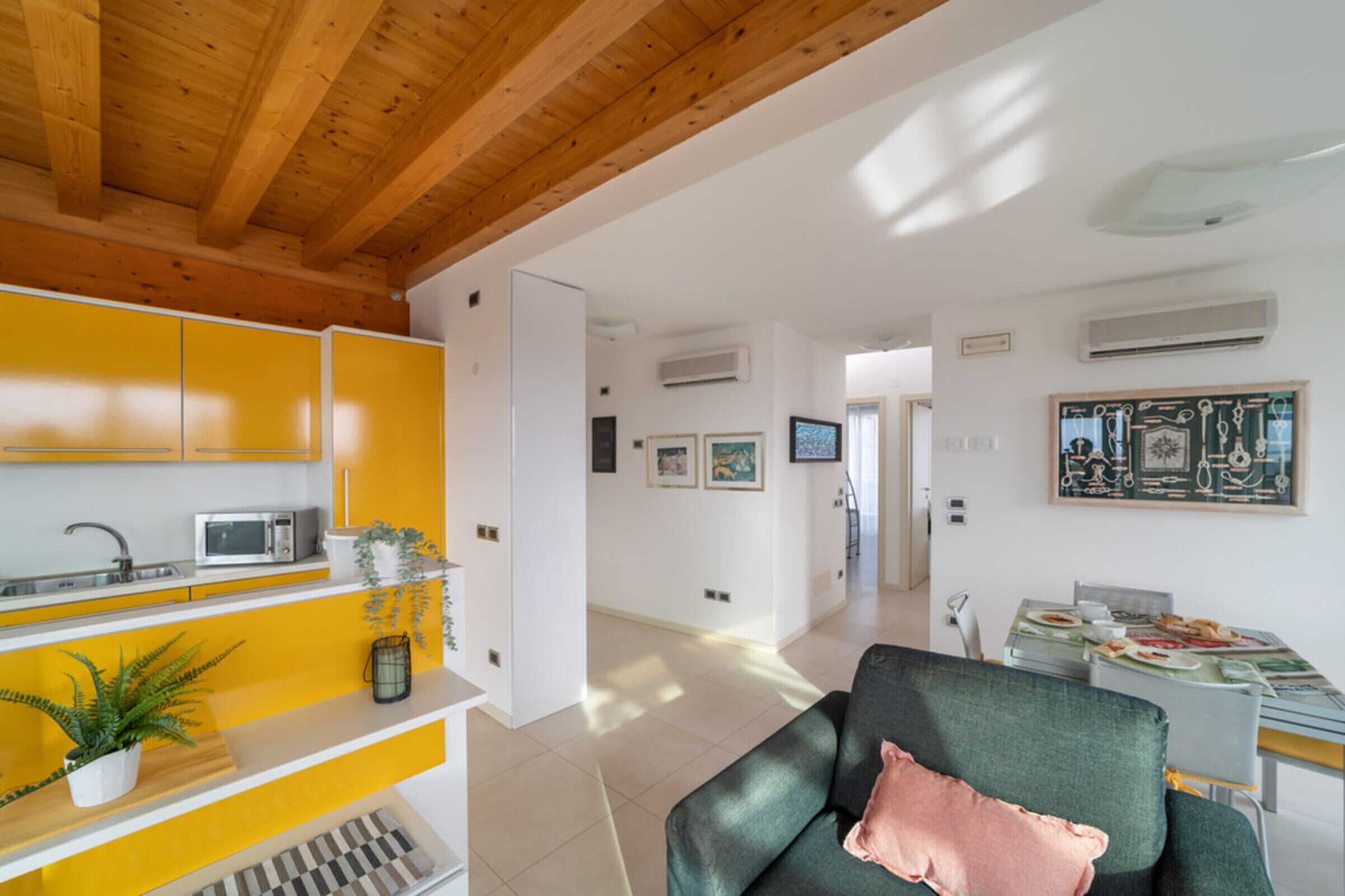 A modern living room with a wooden ceiling, a green sofa, a dining table with a colorful tablecloth, and a kitchen area with yellow cabinets and a microwave.