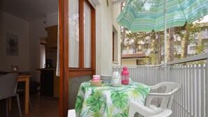 Un espace patio avec une table recouverte d’une nappe à motifs tropicaux, des chaises en plastique blanches, une lanterne rose et un parasol rayé.