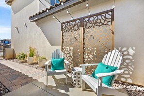 An outdoor seating area with white chairs and a bench, a decorative metal partition, and potted plants. - Desert Escape | Private Fire Pit & Pool Bliss (La Quinta)