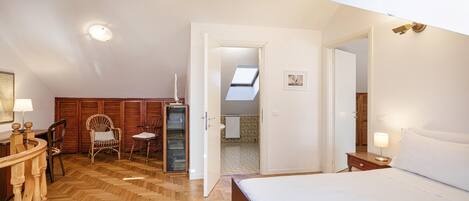 A bedroom with a wooden herringbone floor, a large bed, a desk with a lamp, a chair, and a wardrobe.