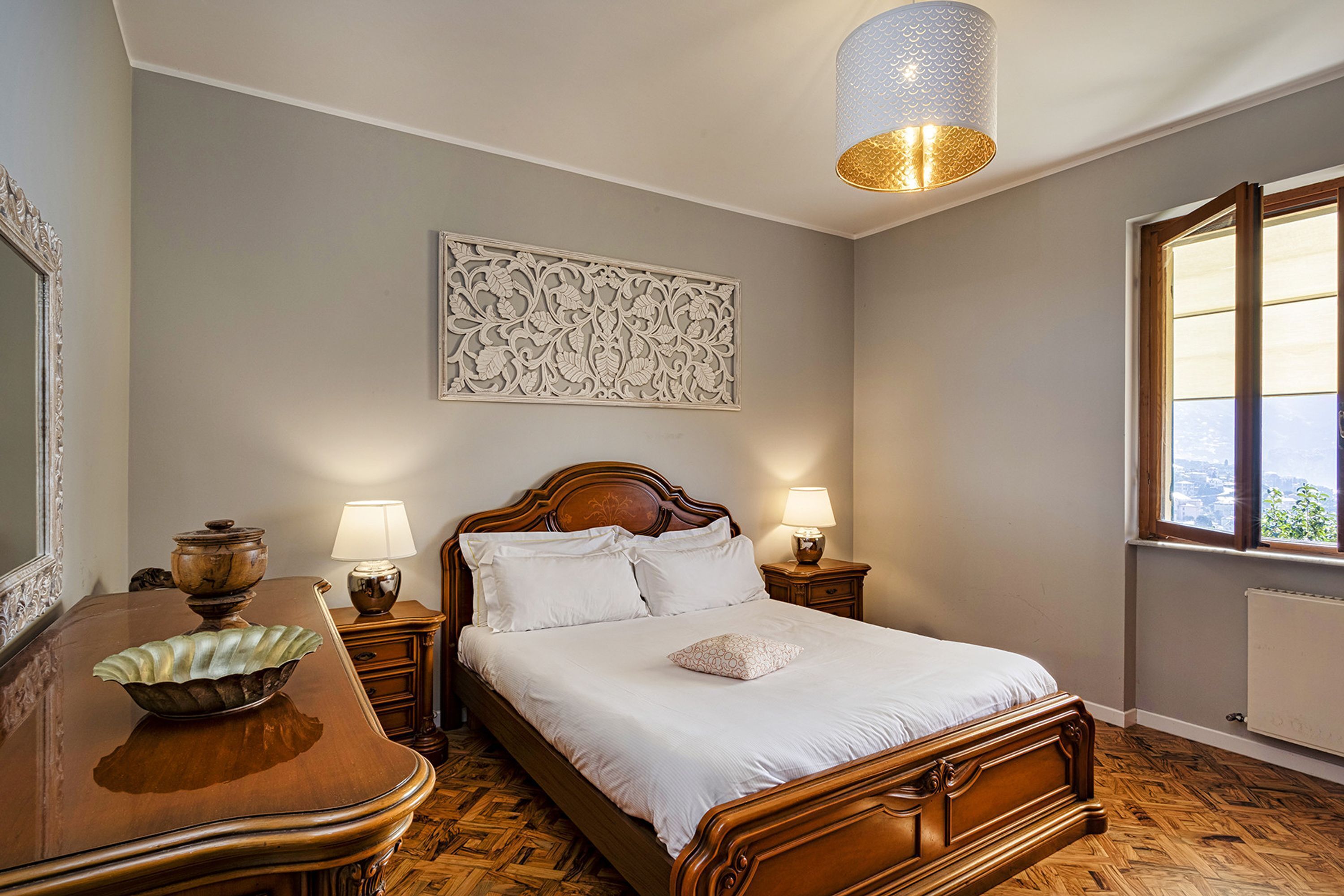 A bedroom with a wooden bed, two bedside tables with lamps, a decorative wall piece, and a window with a view.