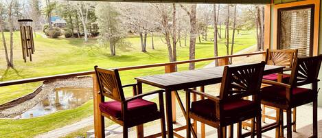A balcony with a wooden table and chairs, a view of a green landscape with trees and a pond.