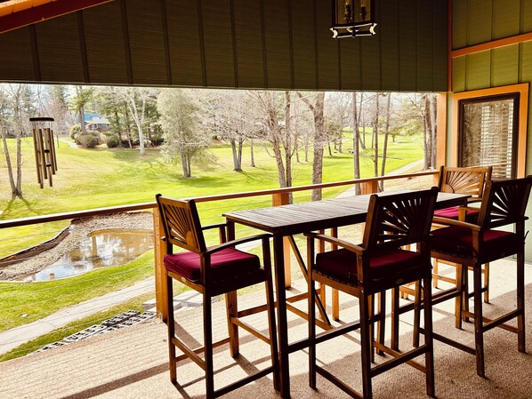 Un balcón con una mesa y sillas de madera, una vista de un paisaje verde con árboles y un estanque.