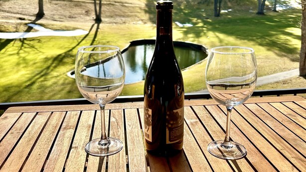 A wooden table with two wine glasses and a bottle of wine, set against a view of a golf course. - NEW Papa's Place on 1st hole Linville Land Harbor Pet Friendly  (Newland)