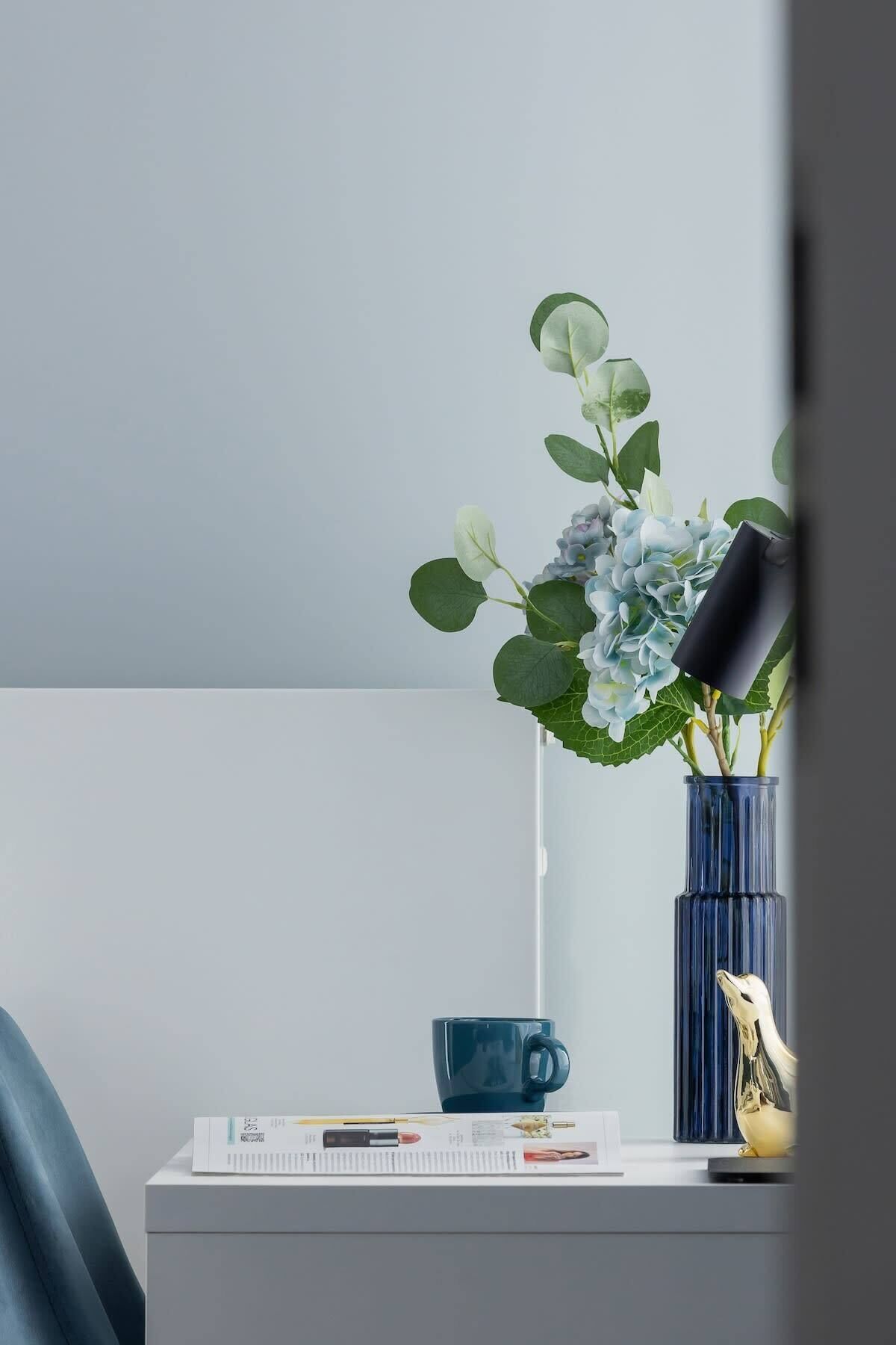 Una scrivania con un vaso di fiori, una tazza blu e una lampada.