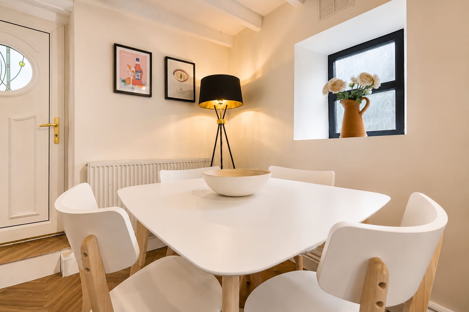 A dining area with a white table, four chairs, a floor lamp, and a window with flowers.
