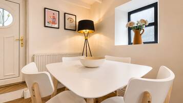 A dining area with a white table, four chairs, a floor lamp, and a window with flowers.