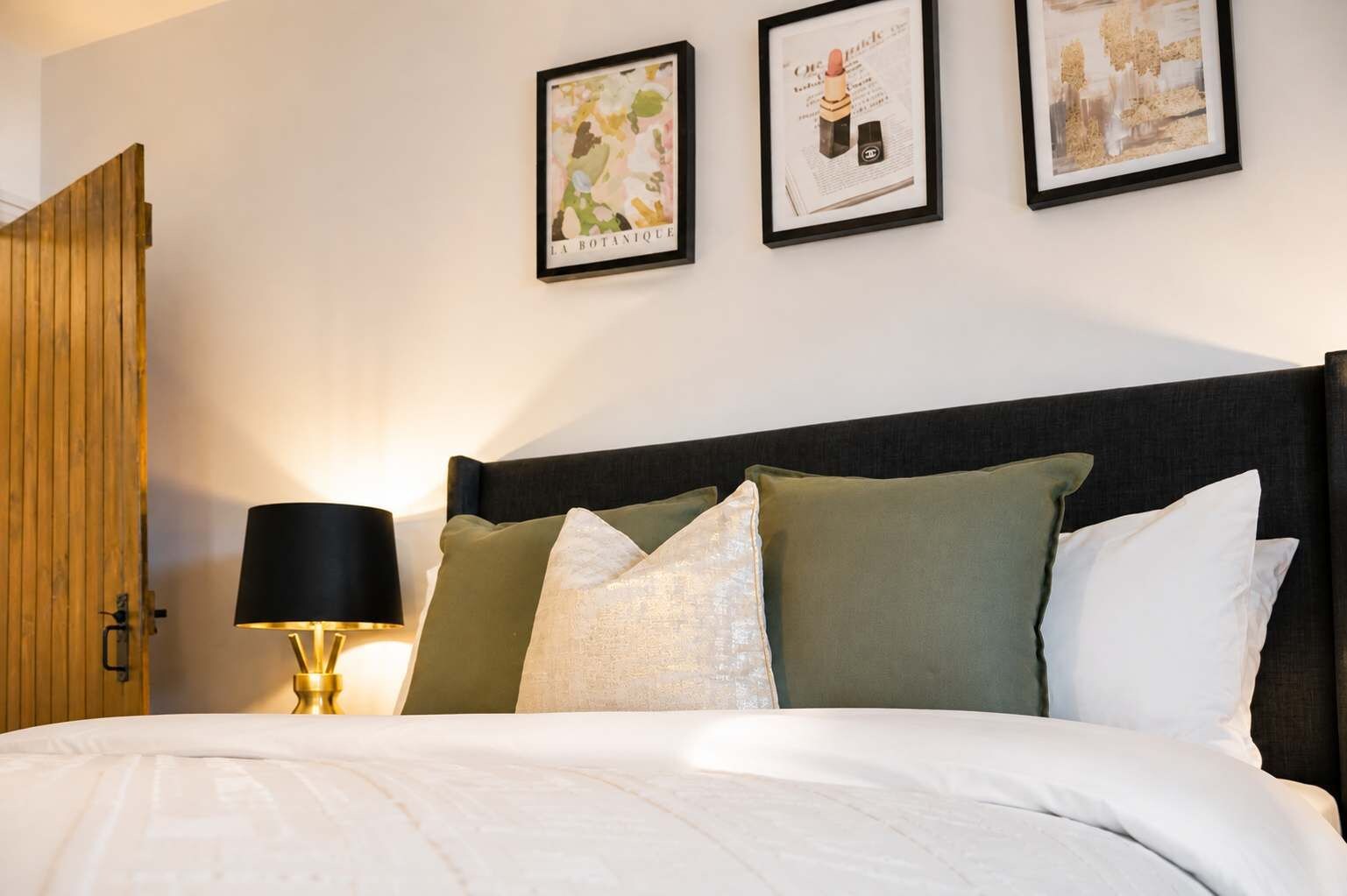 Un lit bien fait, avec une tête de lit sombre, une lampe de chevet et trois tableaux encadrés accrochés au mur.