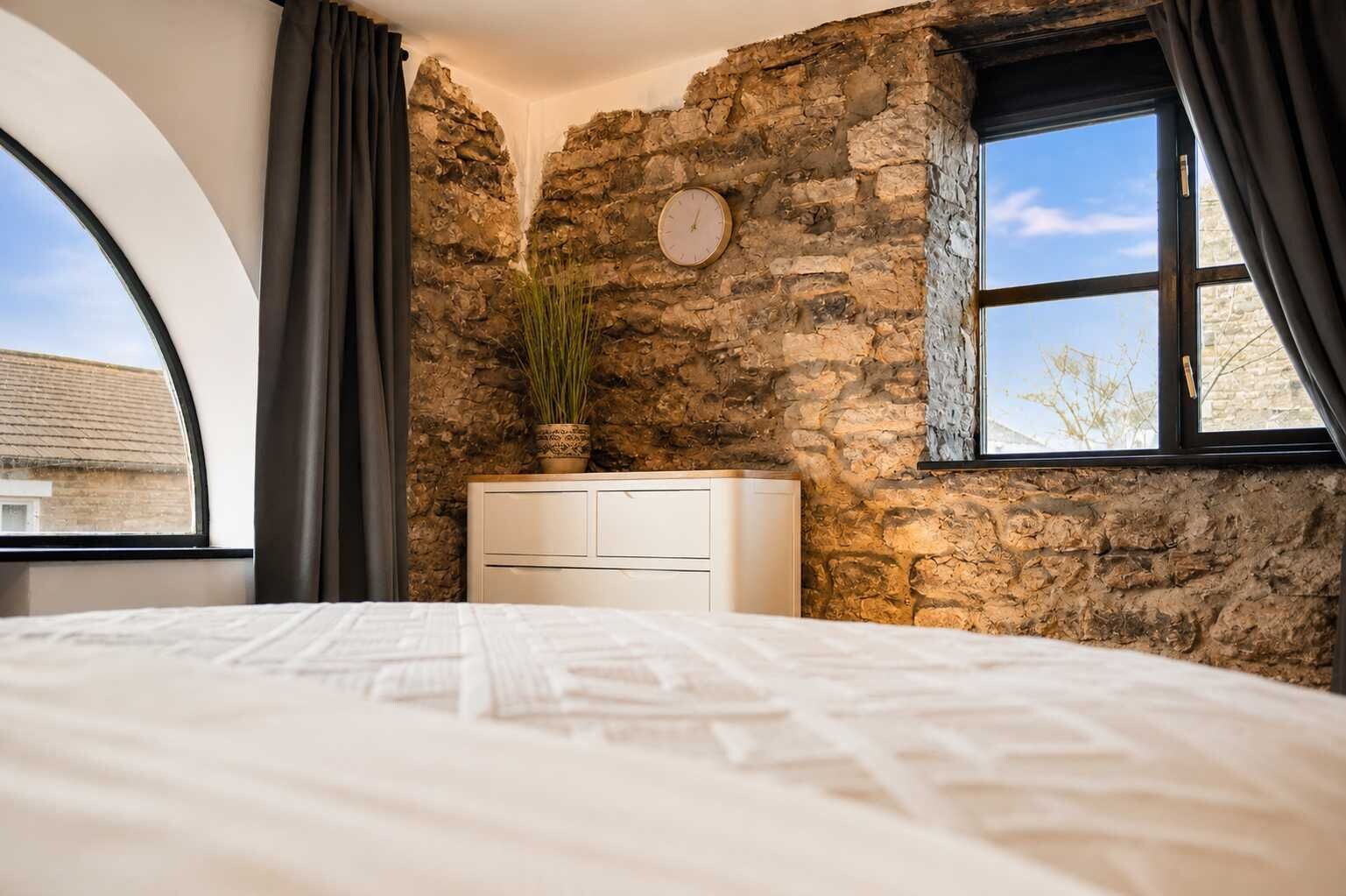 Ein Schlafzimmer mit Steinwand, einem Bett, einem Schminktisch, einer Uhr und einem Fenster mit Vorhängen.
