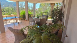 Un patio couvert comprenant une table de ping-pong, des fauteuils en osier, une table à manger avec des chaises et une grande plante en pot.