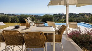 Un área de comedor al aire libre con sillas de mimbre y una mesa, con vistas a una piscina y un paisaje de mar y colinas.