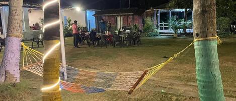 An outdoor area with a hammock, tables, and chairs, surrounded by palm trees.