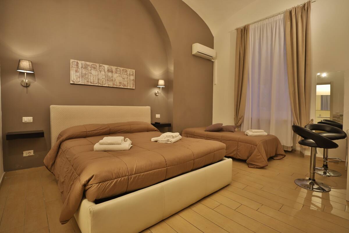 Habitación de hotel con dos camas, una zona de estar con una silla, aire acondicionado de pared y un cuadro en la pared.