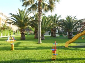 Un parque infantil con columpios y un tobogán, rodeado de palmeras y césped.
