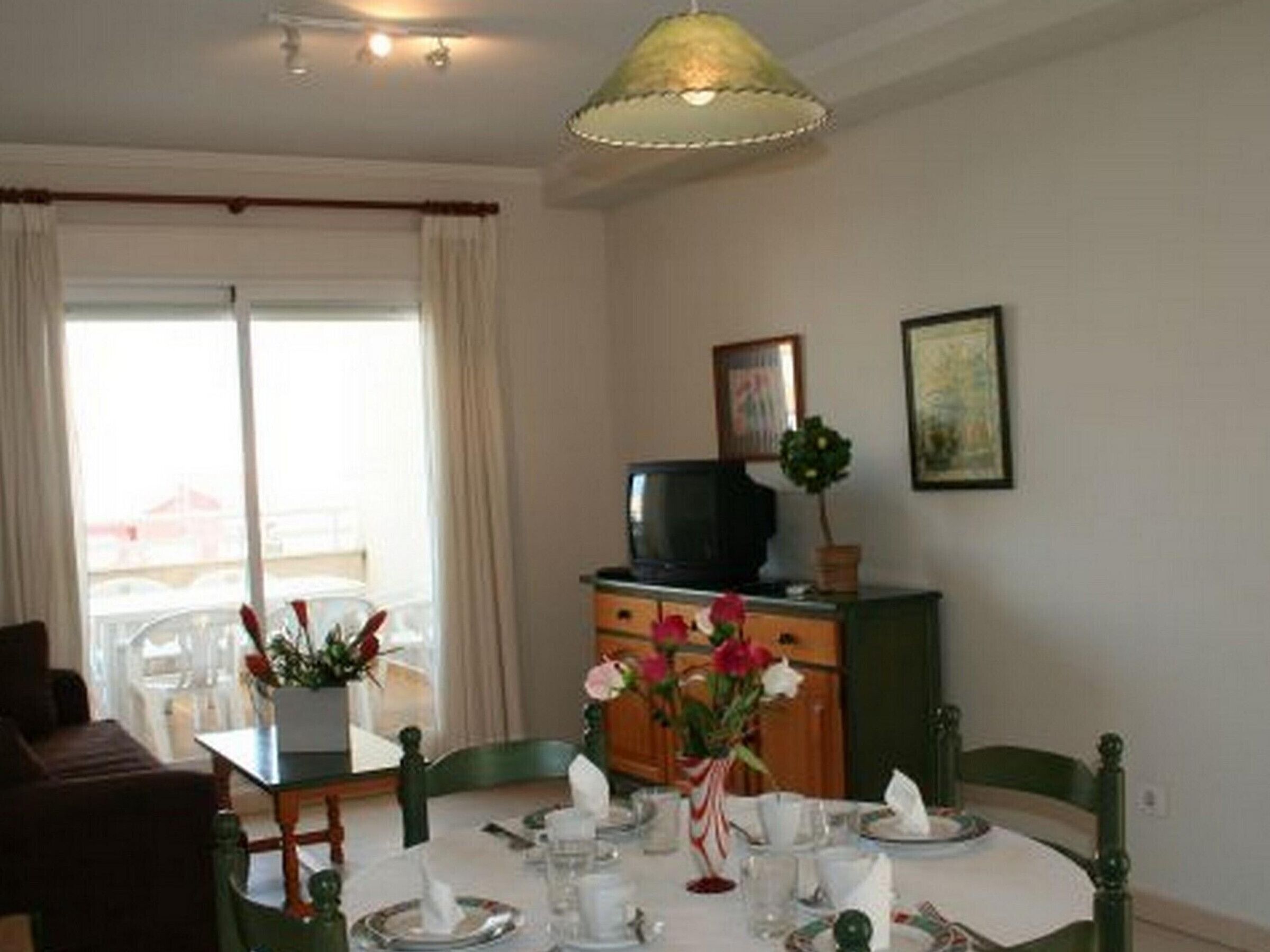A dining area with a round table set for four, a television on a cabinet, and a window with curtains.