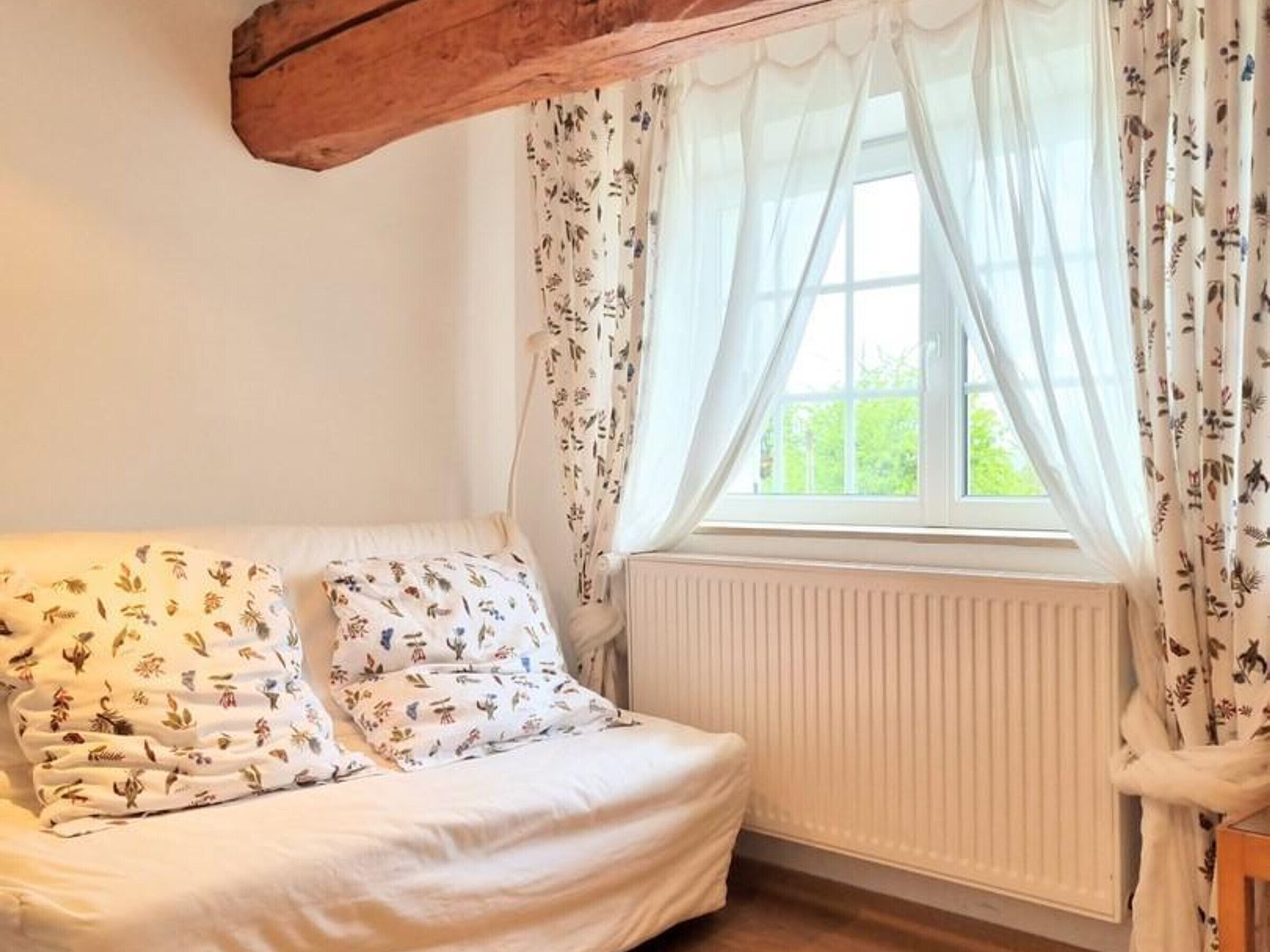 Um quarto aconchegante com uma cama, roupa de cama com padrão floral, uma janela com cortinas leves e um radiador.