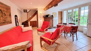 Un salon spacieux avec un canapé rouge, des chaises en bois, une table à manger recouverte d’une nappe rouge, un escalier et de grandes fenêtres donnant sur un paysage verdoyant.